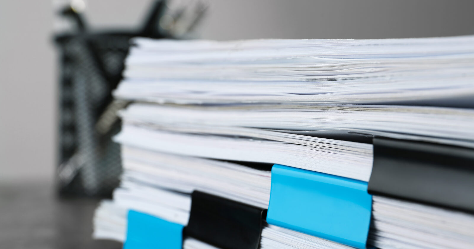 A tall stack of loose white paper held together with blue and black binder clips on a grey desk